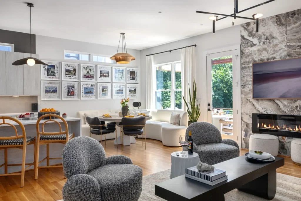 Modern open-concept living room and kitchen captured by an STR photographer, featuring contemporary furniture, a fireplace, wall art, and large windows letting in natural light. Cozy seating and a dining area overlook lush greenery outside.
