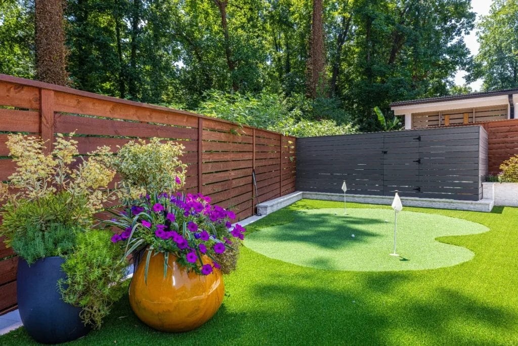 A backyard with artificial grass features a small putting green with two golf flags, bordered by wooden fences and surrounded by large decorative planters, perfect for an str photographer seeking vibrant shots of colorful flowers and lush greenery.