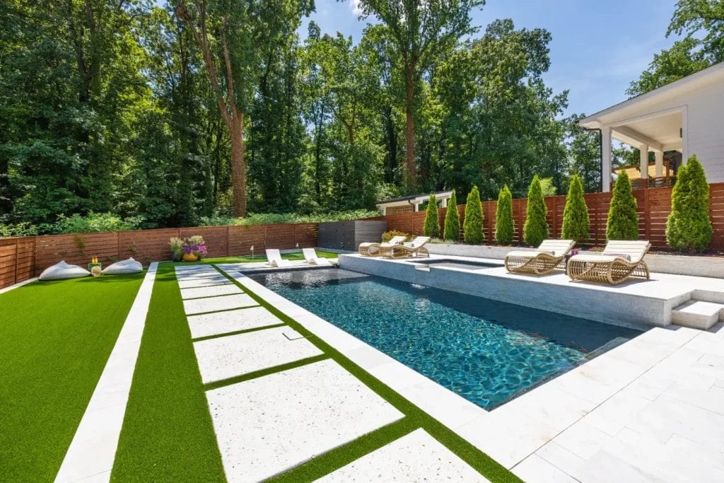 Modern backyard captured by an str photographer with a rectangular pool, lounge chairs, green artificial grass, white stone pathways, and tall trees and shrubs along a wooden privacy fence. A contemporary house is partially visible on the right.