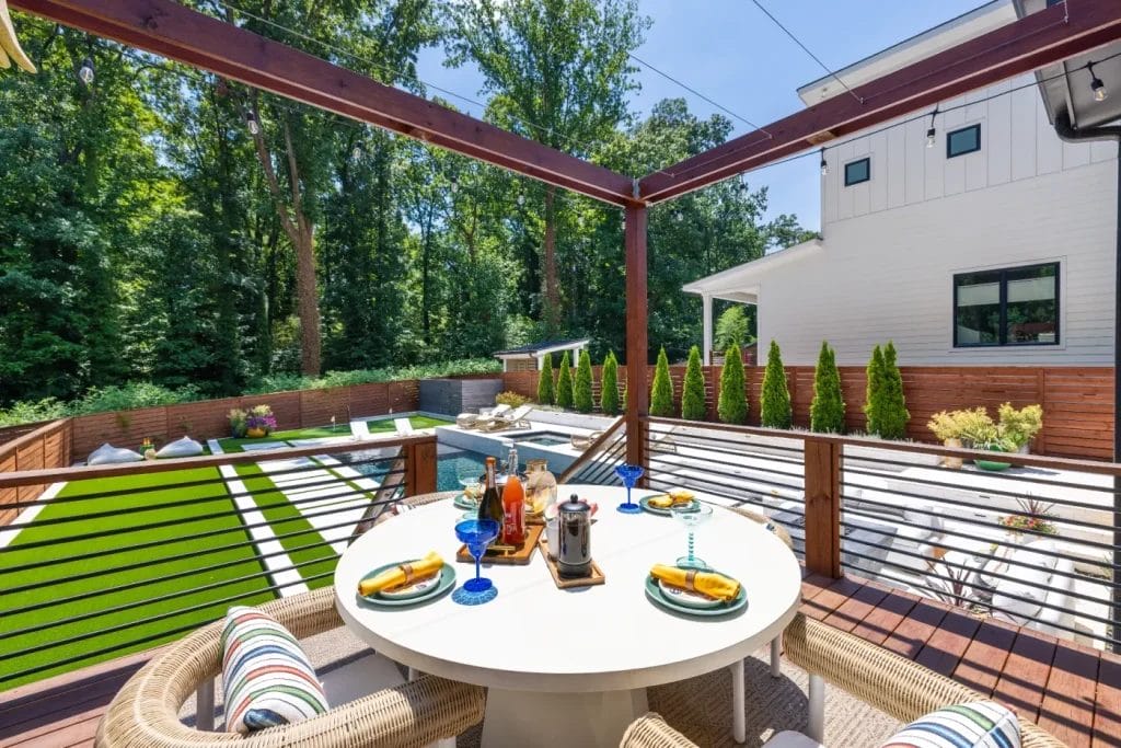 A modern backyard patio beautifully captured by an str photographer, featuring a round dining table for four, overlooking a lawn, pool, and lounge chairs—all bordered by a wooden fence and lush greenery.