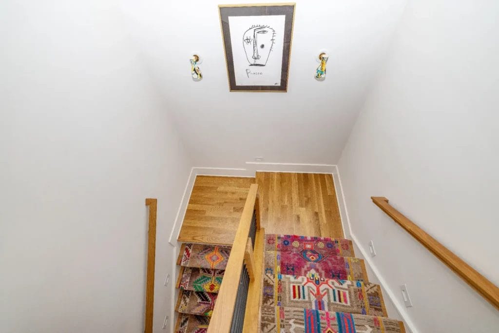 A wooden staircase with colorful, patterned carpet runners leads down to a hardwood landing. A framed abstract face drawing, perfect for any str photographer’s inspiration, hangs on the white wall above the stairs.