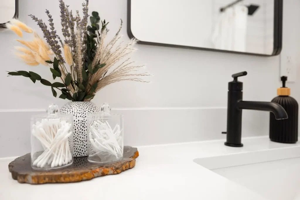 A bathroom countertop styled by an str photographer features two glass jars with cotton swabs and applicators, a vase of dried flowers, a sleek black soap dispenser, and a matching black faucet beside a white sink.