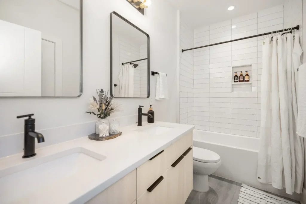 Modern bathroom with double sinks, black fixtures, large mirrors, white countertop, light wood cabinets, and a white tiled bathtub-shower. Perfectly styled for an str photographer seeking bright, clean design and impeccable organization.