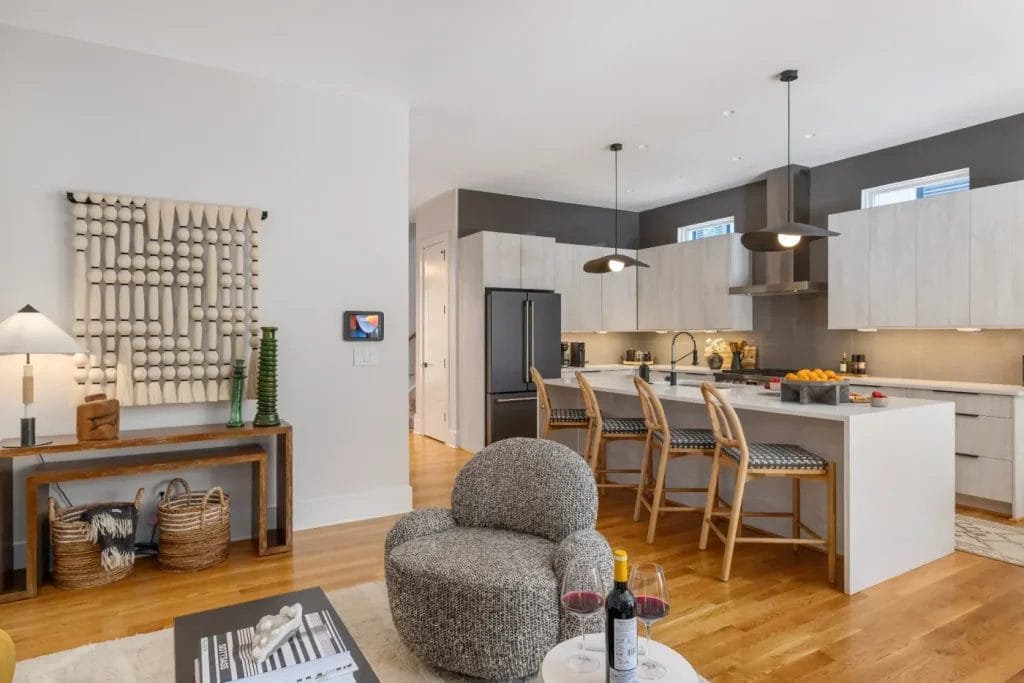 Modern open-concept kitchen and living area perfect for an str photographer, with wooden floors, a cozy gray chair, white island with bar stools, pendant lights, stylish wall art, light cabinets, black fridge, and fresh countertop fruits.