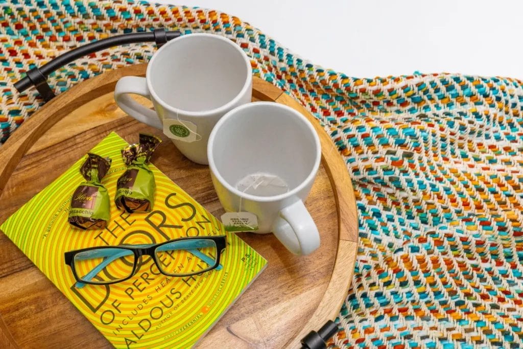 STR photographer’s shot: Two empty white mugs, "The Doors of Perception," a pair of glasses, and two wrapped chocolates rest on a wooden tray beside a colorful woven blanket.