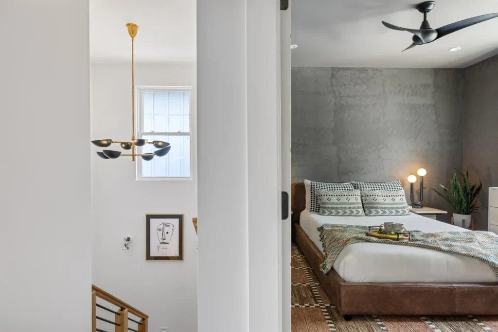 Modern bedroom with a brown bed, patterned bedding, and gray accent wall; str photographer captures the adjacent hallway’s gold chandelier, framed art, and window, separated by a white wall and sliding door.