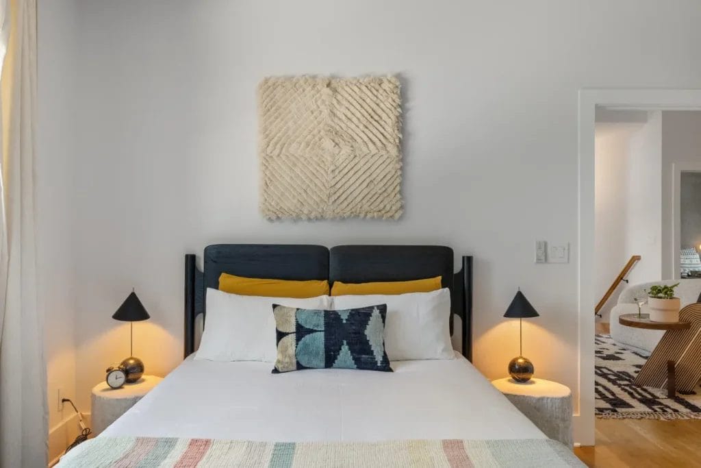 A modern bedroom with a dark bedframe, white bedding, and a patterned pillow is beautifully captured by an str photographer. Two round nightstands with matching black lamps and alarm clocks flank the bed, while a textured wall hanging decorates the light gray wall.