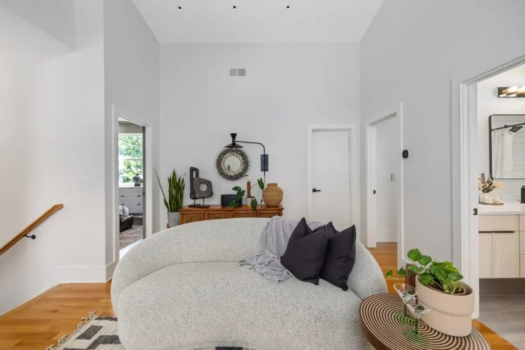 A modern living room, perfect for a str photographer, features a curved white sofa with black and gray pillows, a round side table with plants and a glass, wooden floors, plus a console table with decor and a round mirror set against a white wall.
