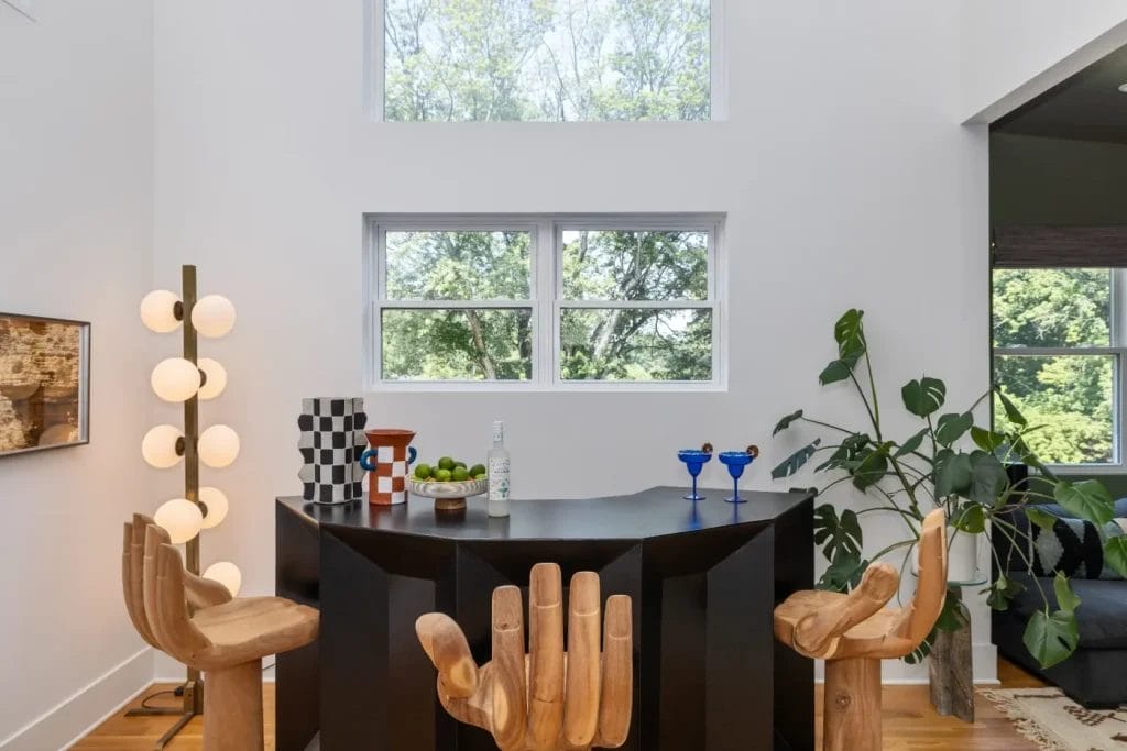 A modern home bar with a black geometric counter, two hand-shaped wooden chairs, and various glassware. Large windows let in natural light, ideal for an str photographer capturing interiors. A potted plant and unique floor lamp complete the scene.