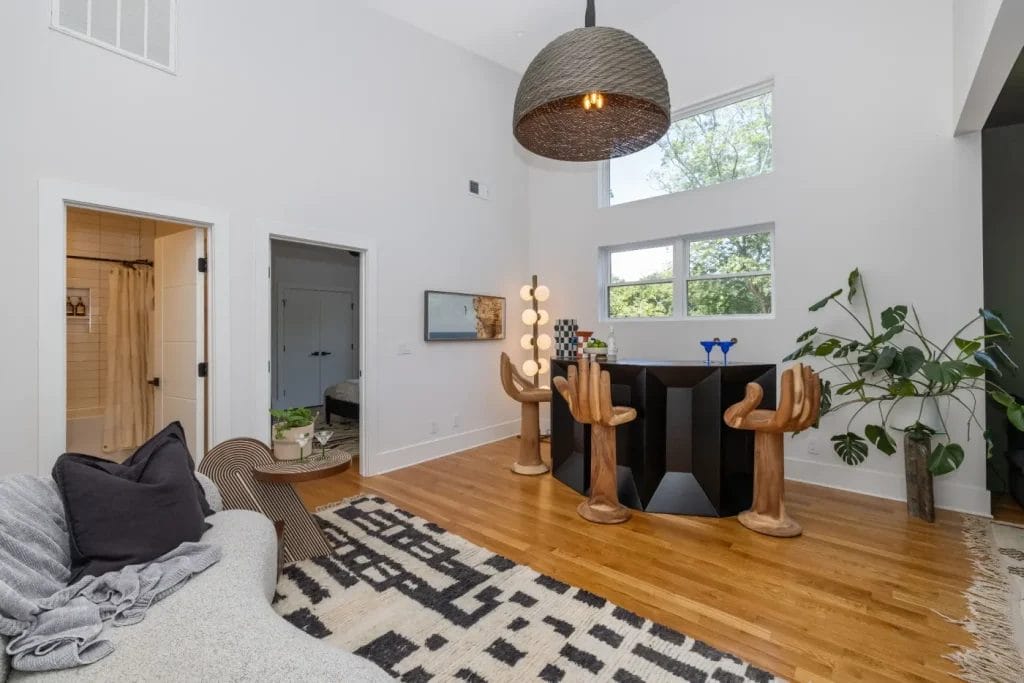 Modern living room with wood floors, a geometric black-and-white rug, a bar area featuring unique hand-shaped chairs, large windows perfect for any str photographer, a gray sofa, potted plants, and a woven pendant light hanging from the ceiling.