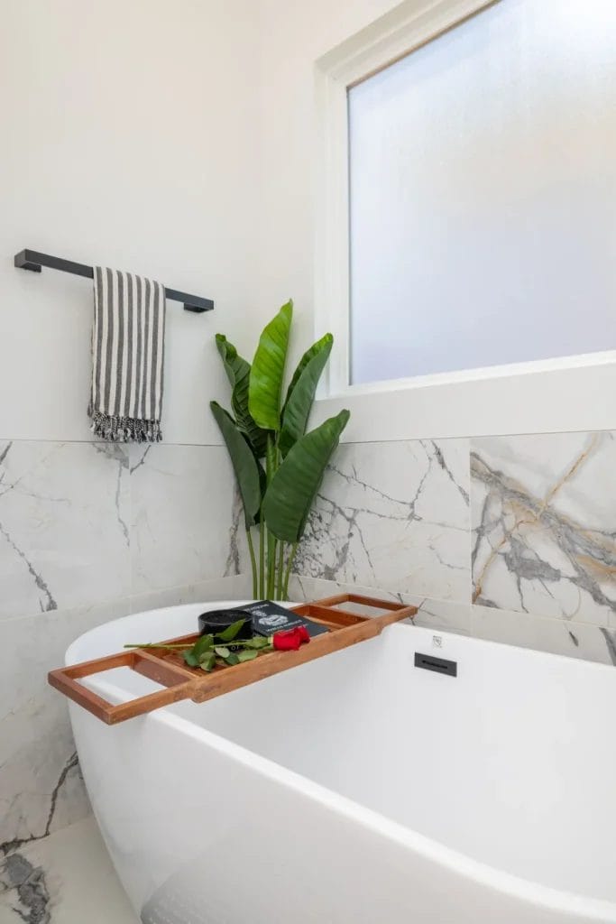 A modern white bathtub is styled by a str photographer, featuring a wooden tray with a book, green leaves, and a red rose. Marble walls, a tall plant, frosted window, and striped towel on a black rack complete this serene space.