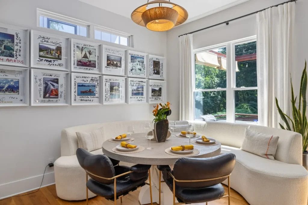 A modern dining nook, ideal for an str photographer, features a round table set for four, white curved banquette seating, black chairs, and large windows. Artworks adorn the wall beneath a contemporary pendant lamp above the table.