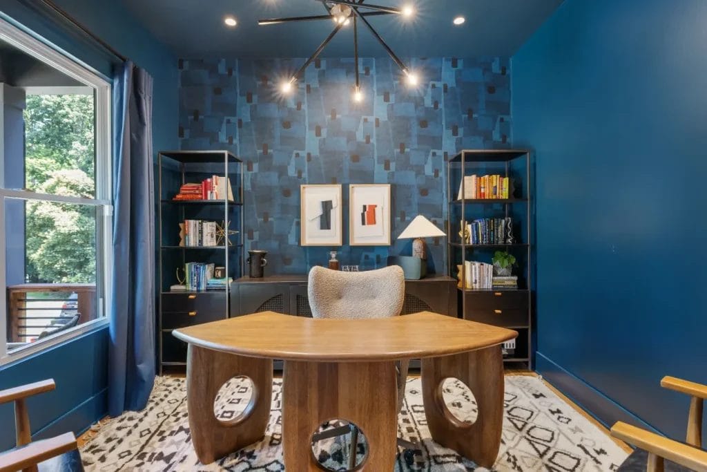 A stylish home office with deep blue walls, geometric wallpaper, a modern wood desk, and cozy chair—perfect for a str photographer. Bookshelves, abstract art, and a patterned rug complete the space as sunlight streams through a large window on the left.