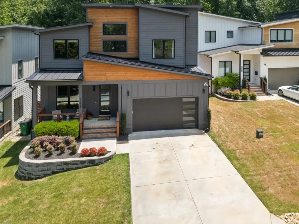 Modern two-story house with gray siding, wood accents, large windows, and a covered front porch—ideal for an STR photographer to capture. Features attached garage, well-kept lawn, landscaped flower bed, and neighboring houses on both sides.