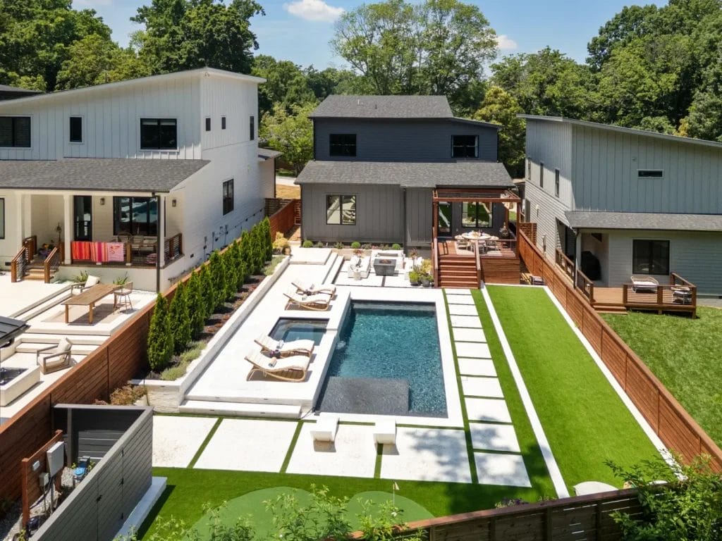 A modern backyard captured by an str photographer showcases a rectangular pool, lounge chairs, green lawn, and two contemporary houses separated by wooden fences and surrounded by trees on a sunny day.