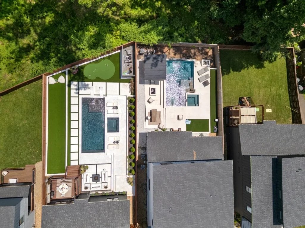 Aerial view by str photographer shows two modern backyards—each with a rectangular swimming pool, lounge areas, and landscaped greenery. One yard features a putting green; both fenced and adjacent to large gray-roofed houses.