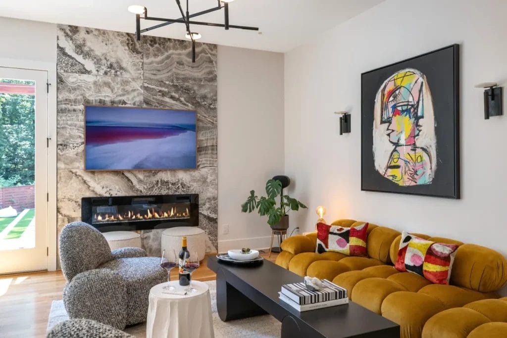 A modern living room styled for an str photographer, featuring a mustard yellow tufted sofa, patterned cushions, abstract wall art, a black coffee table, mounted TV above a marble fireplace, and a round chair by the window.