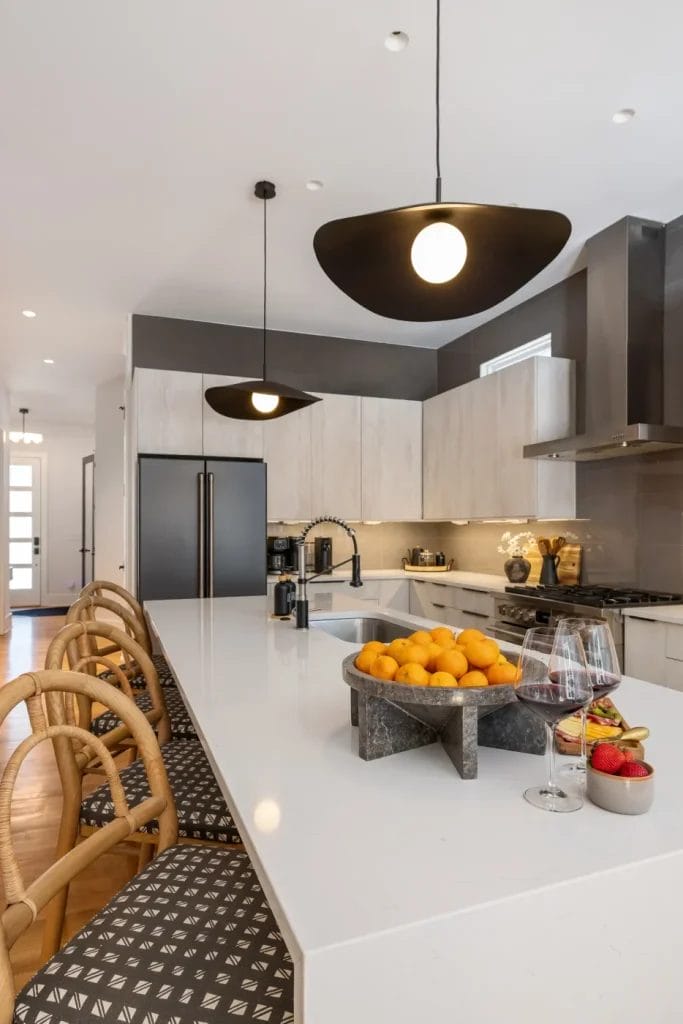 Modern kitchen with a white island countertop captured by an str photographer. Features rattan chairs, pendant lights, a bowl of oranges, a glass of wine, and strawberries. Light wood cabinets, black refrigerator, and stainless steel appliances in the background.