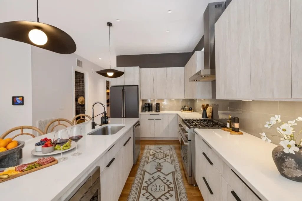 Modern kitchen with light wood cabinets, white countertops, stainless steel appliances, and pendant lights. Captured by an str photographer, the space features a central island with fruit, wine, a charcuterie board, rug, and flowers.