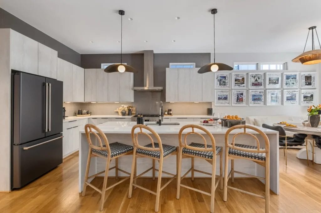 Modern kitchen with light wood cabinets, black refrigerator, and a central island with four rattan bar stools. Captured by an STR photographer, pendant lights hang above the island and a dining area is visible to the right.