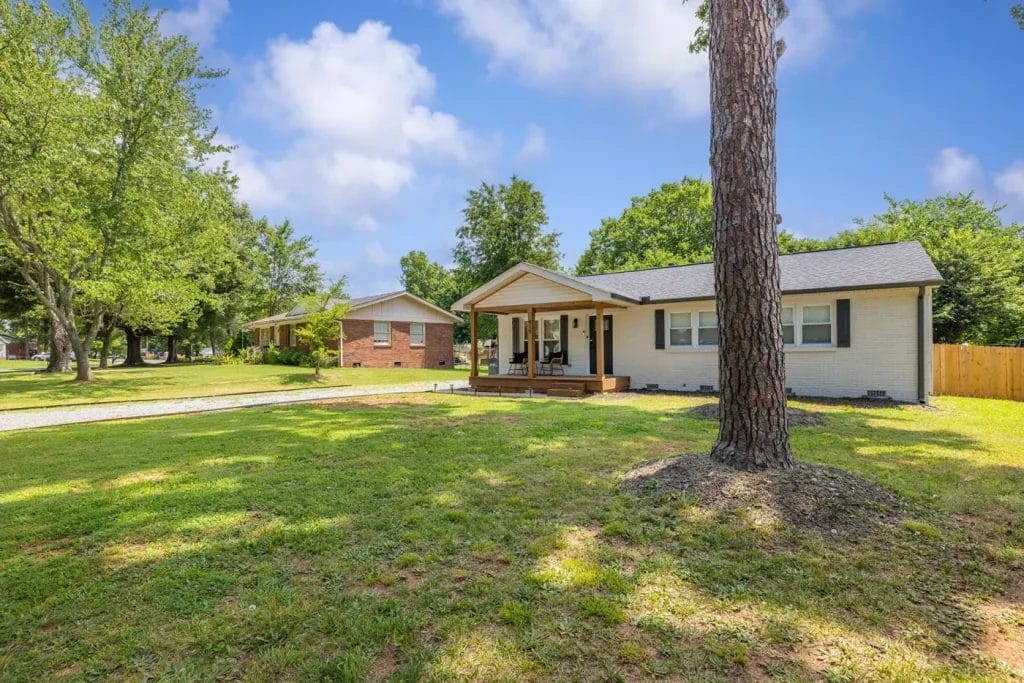 Single-story house with a covered front porch, gray siding, and a dark roof, set on a spacious grassy lawn with trees, a driveway, and a neighboring brick house under a partly cloudy sky.
