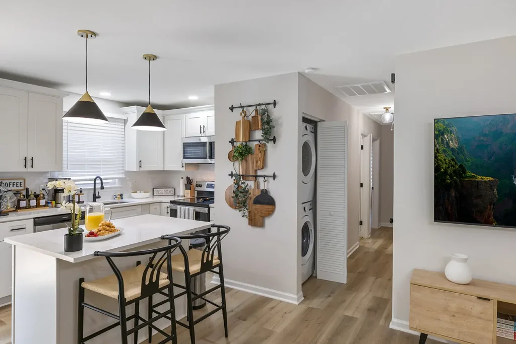 A modern kitchen with white cabinets, a center island with two chairs, pendant lights, and a tray of juice and pastries. A stacked washer and dryer are in an alcove, and a hallway leads to other rooms.