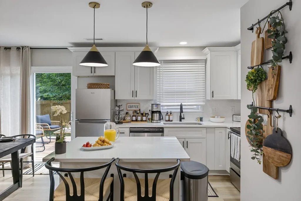 Modern kitchen with white cabinets, stainless steel appliances, and a center island set with breakfast foods, juice, and fruit. Black pendant lights hang above, and a rack with cutting boards decorates the wall. Patio visible outside.