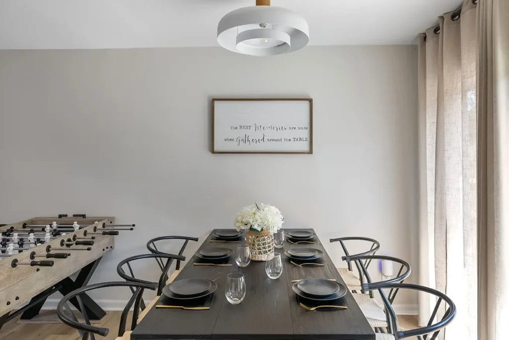 A modern dining room with a dark wooden table set for six, black chairs, a vase of white flowers, and a foosball table on the left. Neutral walls, beige curtains, and a framed quote complete the décor.