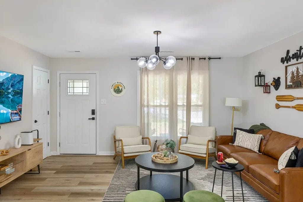 A cozy living room with a brown leather sofa, two beige armchairs, a round coffee table, wall art, a TV, and large windows with sheer curtains letting in natural light.