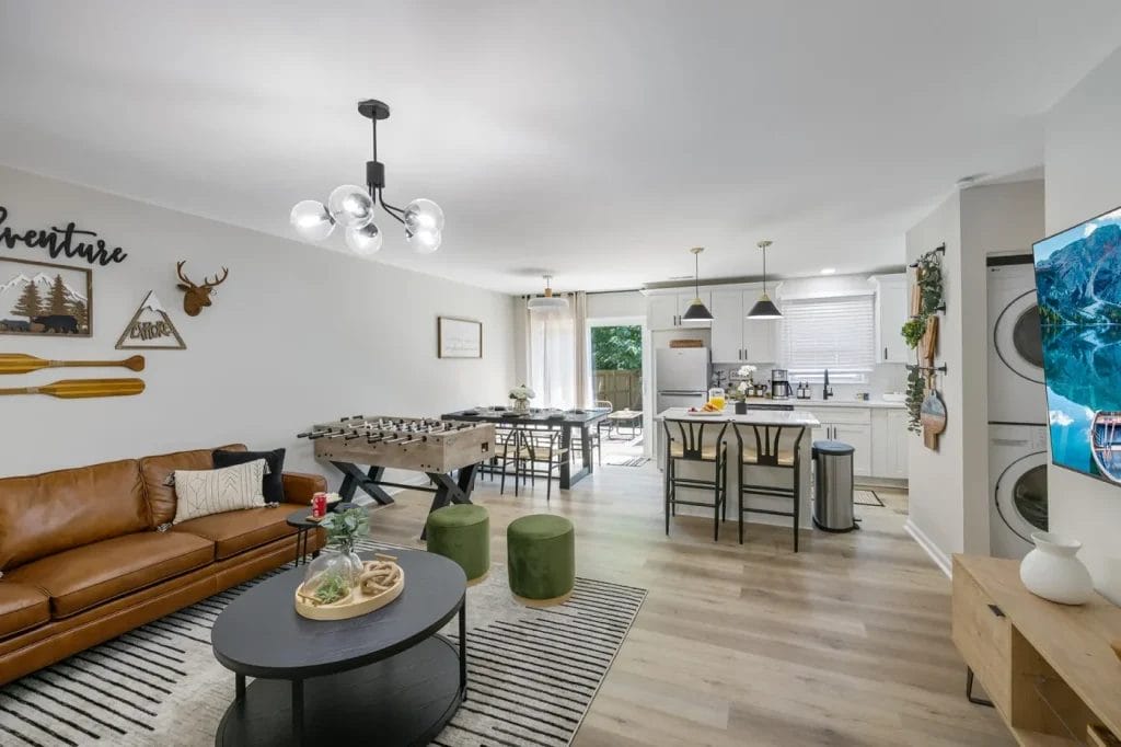 Modern open-concept living room and kitchen, featuring a brown leather sofa, foosball table, wall decor, round coffee table, striped rug, and green stools. The kitchen has white cabinets, pendant lights, and stacked washer-dryer.