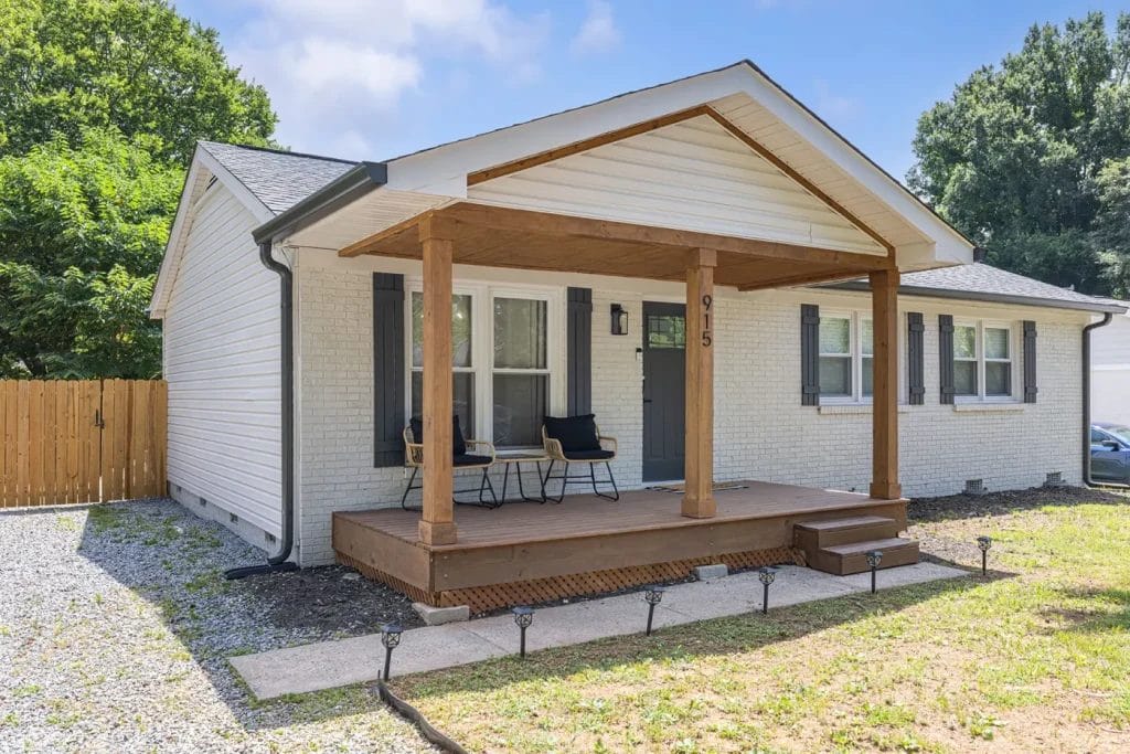 A single-story white brick house with a wooden front porch, two chairs, and a small table. The house has dark shutters, a gray front door, and is surrounded by a grassy yard with trees and a wooden fence.