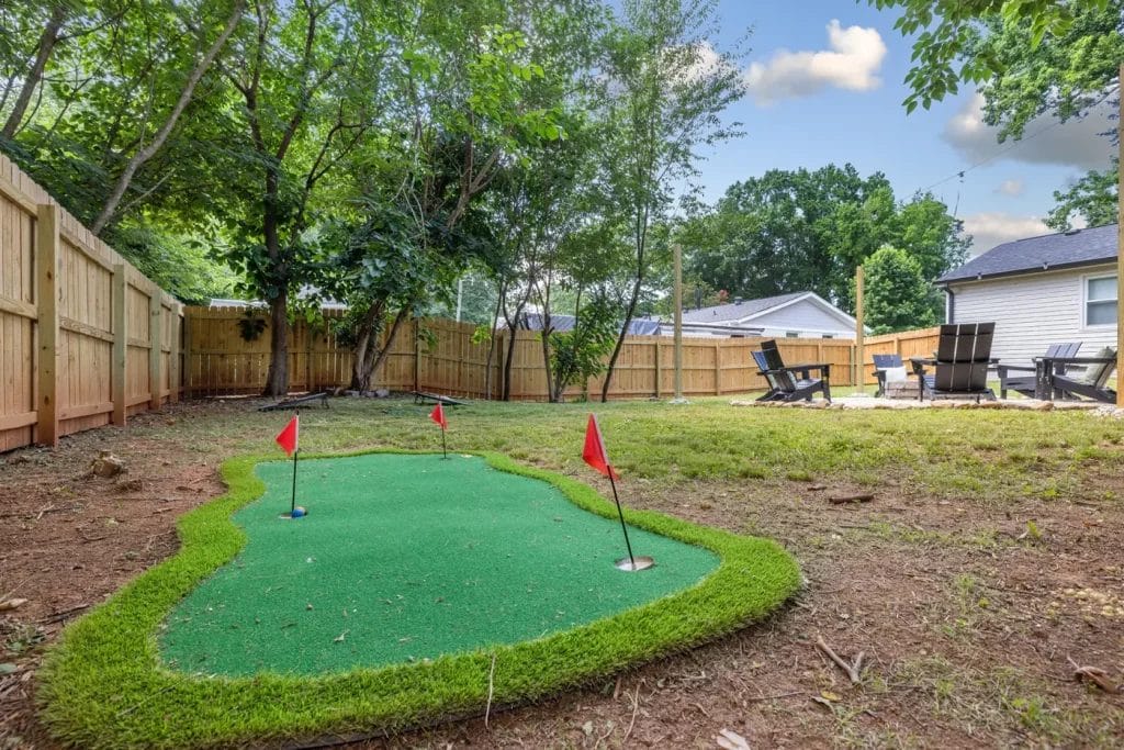 A backyard with a small artificial putting green featuring three red flags, surrounded by grass and trees, and a wooden fence. Patio chairs are visible in the background near a house.