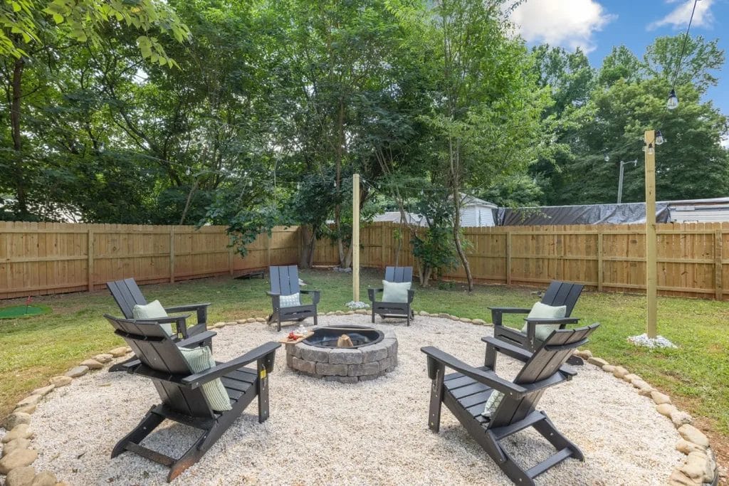 A circular outdoor fire pit area with six black Adirondack chairs arranged around a stone fire pit on a gravel surface, surrounded by a fenced backyard with trees and string lights above.