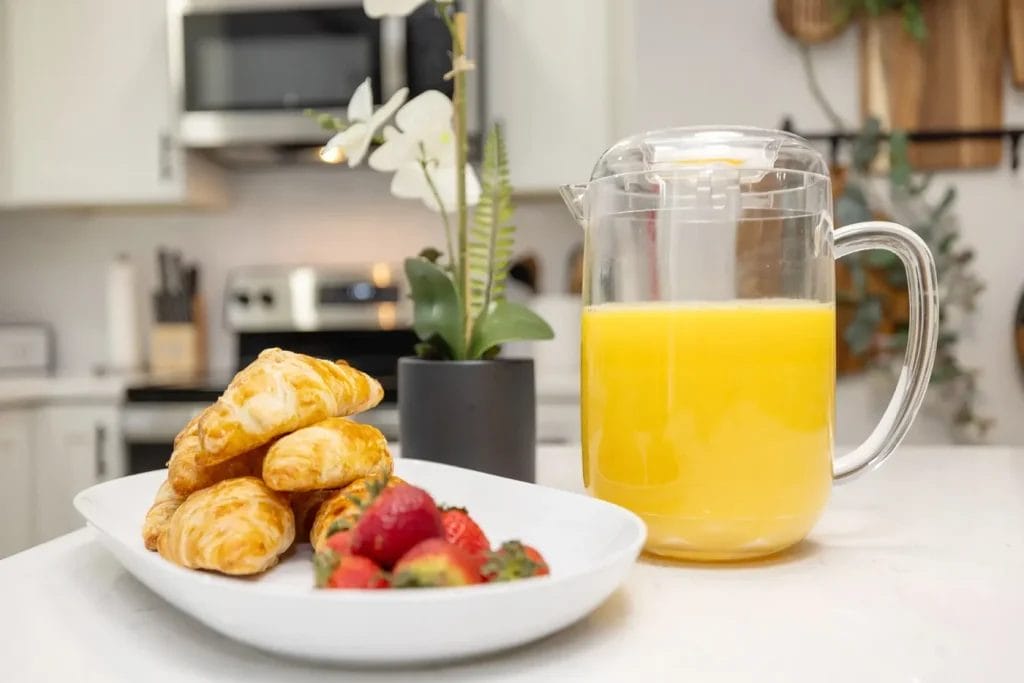 A plate with croissants and strawberries sits on a white counter beside a clear pitcher of orange juice. A small potted plant is in the background, with a kitchen visible behind it.