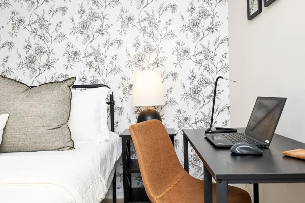 A cozy bedroom workspace featuring a brown chair at a black desk with a laptop and mouse, next to a bed with white bedding and pillows. Behind, a floral-patterned wallpaper and a lit lamp add a decorative touch.