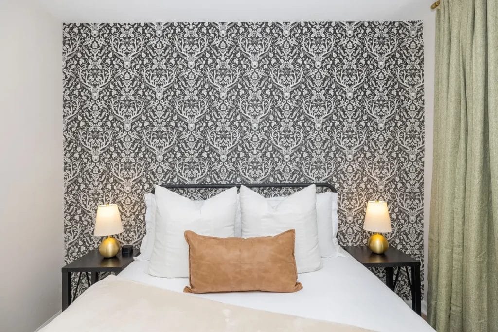 A neatly made bed with white pillows and a tan accent pillow stands against a black and white damask-patterned wallpaper. There are two bedside tables, each with a gold lamp, and green curtains visible on the right.