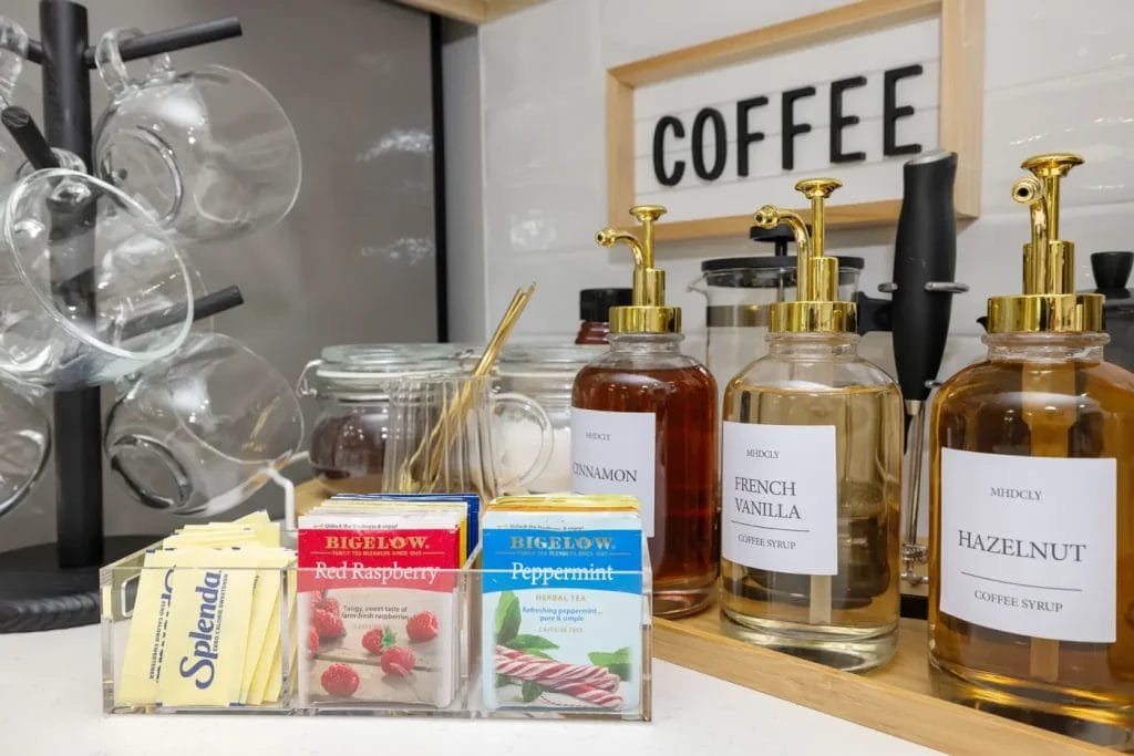 A coffee station with glass syrup bottles labeled cinnamon, French vanilla, and hazelnut, tea bags, packets of Splenda sweetener, mugs on a rack, stirrers in a jar, and a sign that says "COFFEE.