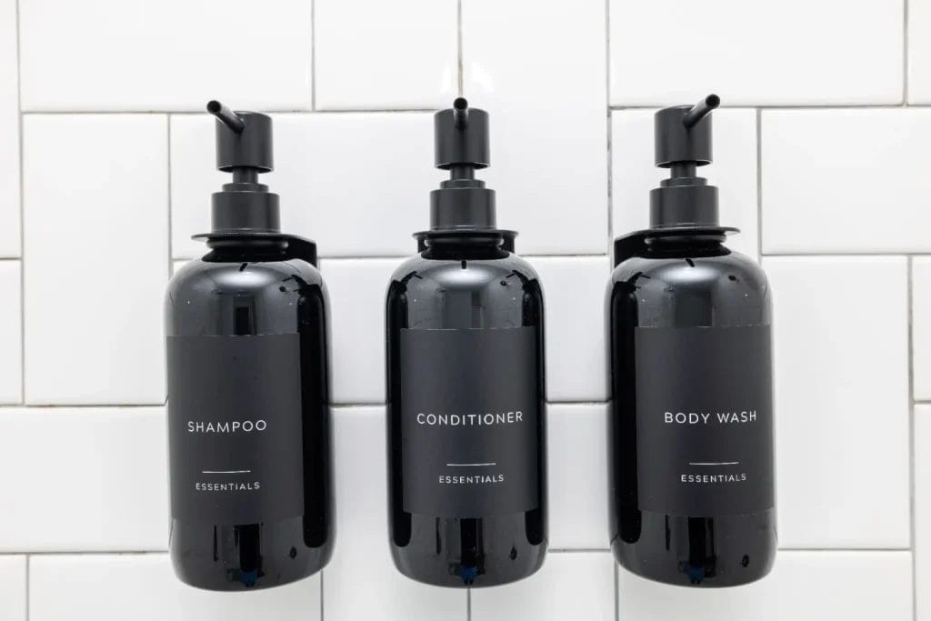 Three black pump bottles labeled shampoo, conditioner, and body wash are mounted on a white tiled wall. Each bottle has simple, minimalist labels with the word "ESSENTIALS" printed underneath.