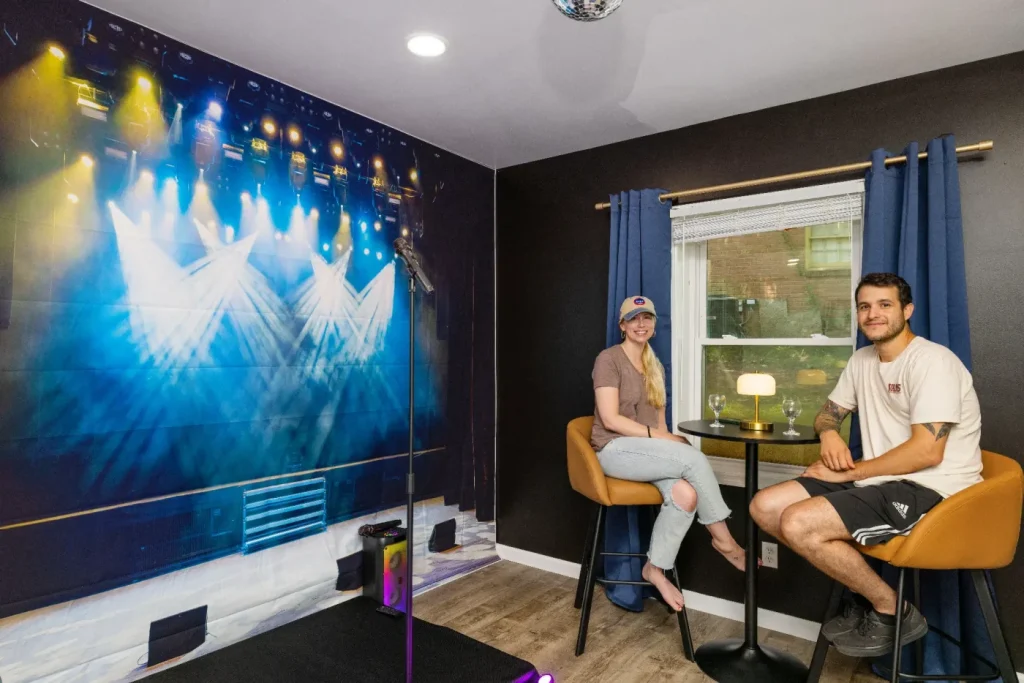 Two people sit at a small round table by a window in a room with a stage-themed wall mural, bar stools, blue curtains, and a microphone stand on a small platform. Warm lighting creates a cozy atmosphere.