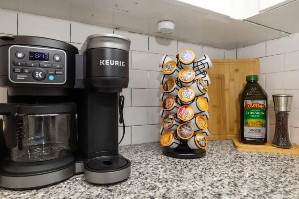 A Keurig coffee maker sits on a kitchen counter next to a rotating stand filled with coffee pods. Nearby are a bottle of Pompeian olive oil, a pepper grinder, and a wooden cutting board against a tiled backsplash.