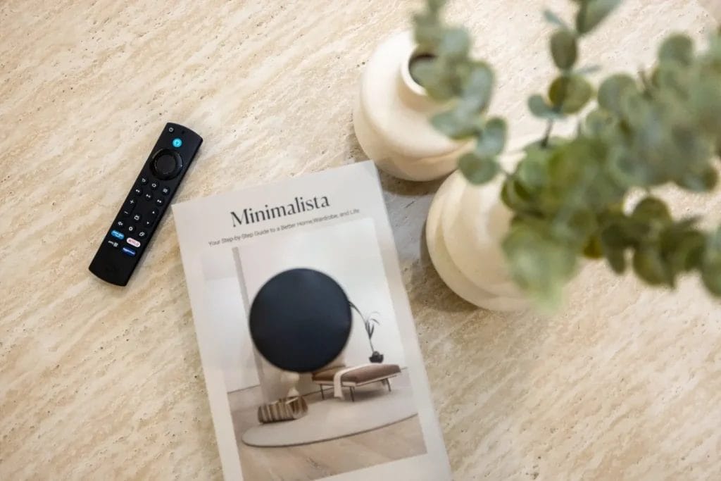 A book titled "Minimalista" lies on a light-colored table next to a TV remote and two white vases with green foliage. The scene has a calm, minimalist aesthetic.