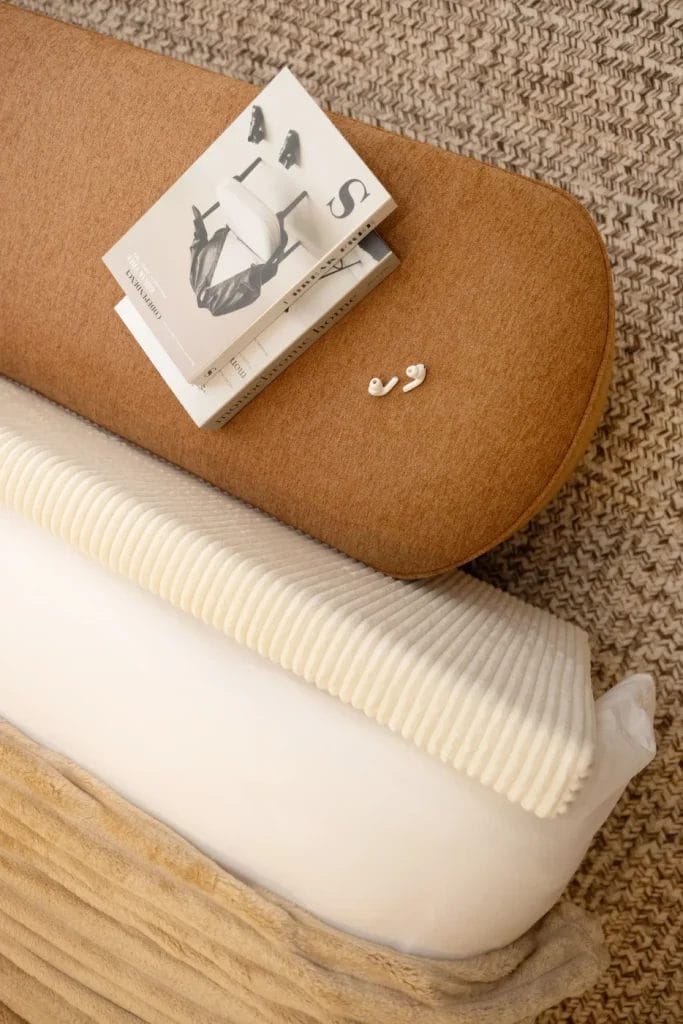 A stack of magazines, a pair of gold hoop earrings, and a book rest on a brown cushioned bench above a white, ribbed blanket and textured beige throw, all placed on a woven rug.