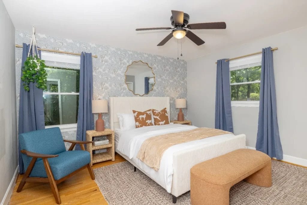 A bright bedroom with a white bed, tan throw, animal-print pillows, round mirror, two bedside tables with lamps, blue curtains, a blue armchair, a tan bench, a ceiling fan, and a potted plant.