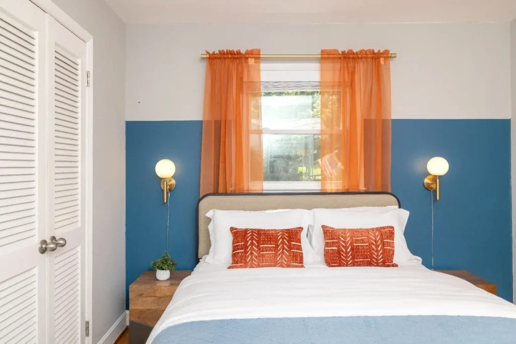 A neatly made bed with white linens, orange decorative pillows, and a blue blanket sits against a blue and gray wall. An orange sheer curtain hangs over a window above the bed, flanked by two wall-mounted globe lights.