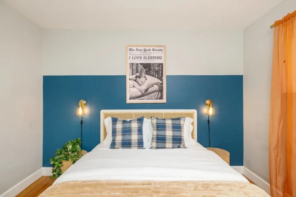 A modern bedroom with a bed against a blue and gray accent wall, plaid pillows, a beige blanket, two small side tables with lamps, a potted plant, orange curtains, and a framed poster above the bed.