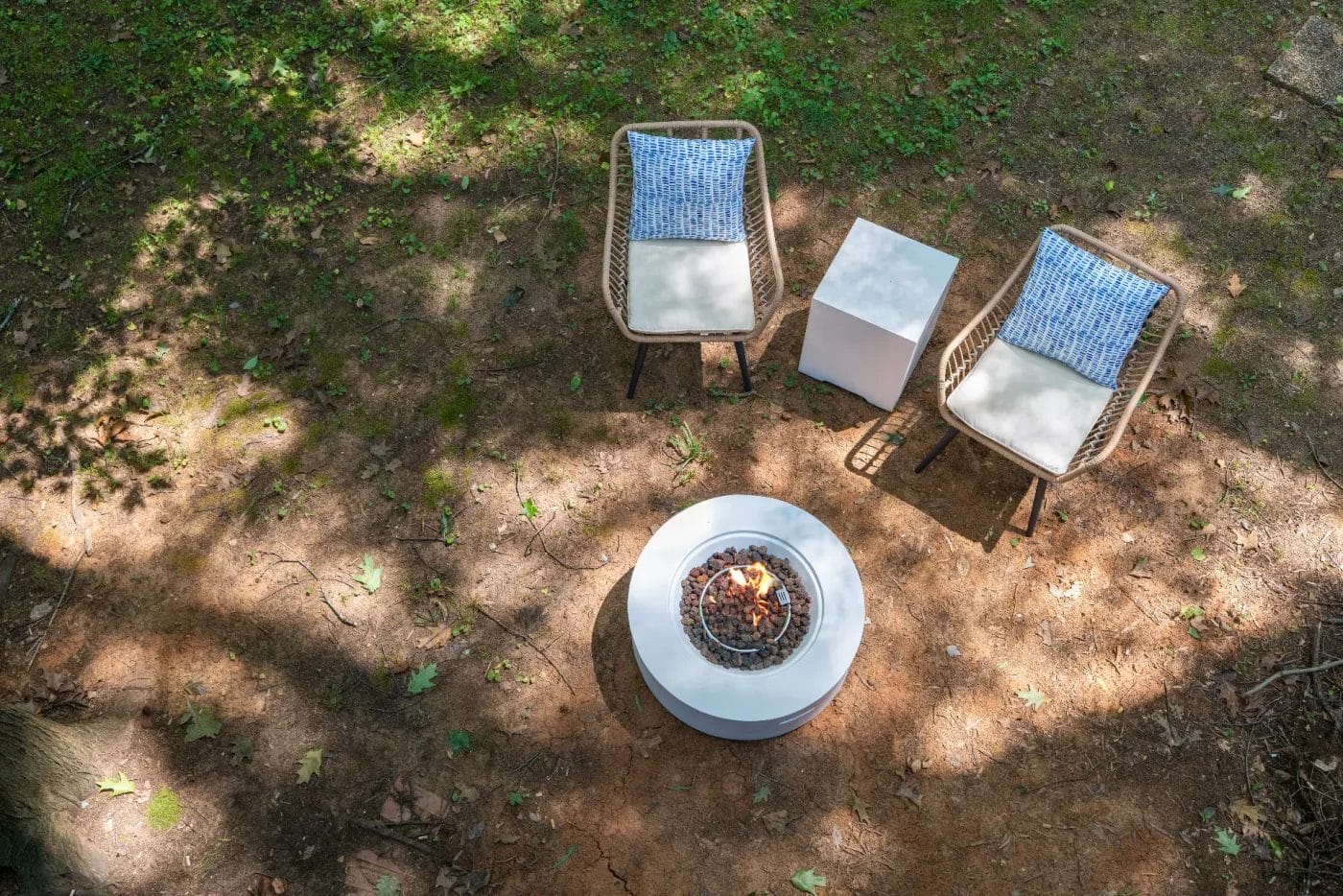 Aerial view of two wicker chairs with blue-patterned cushions, a white square side table, and a round white fire pit with a small fire, set on a patch of sun-dappled dirt and grass.