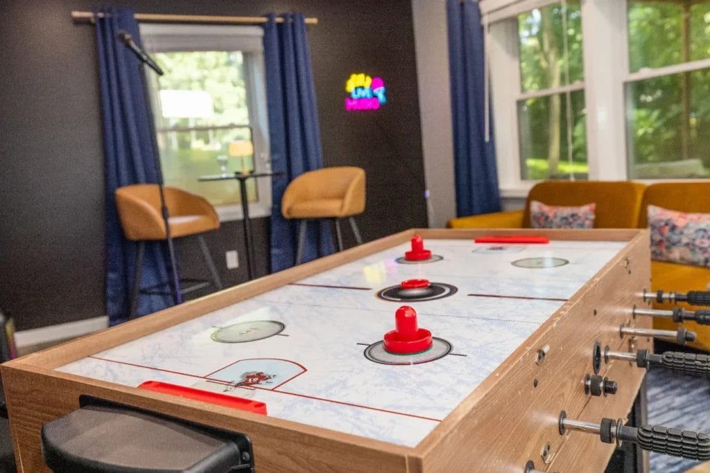 A close-up of an air hockey table in a cozy game room with blue curtains, yellow chairs, a microphone stand, and a neon "Game Room" sign near large windows letting in natural light.