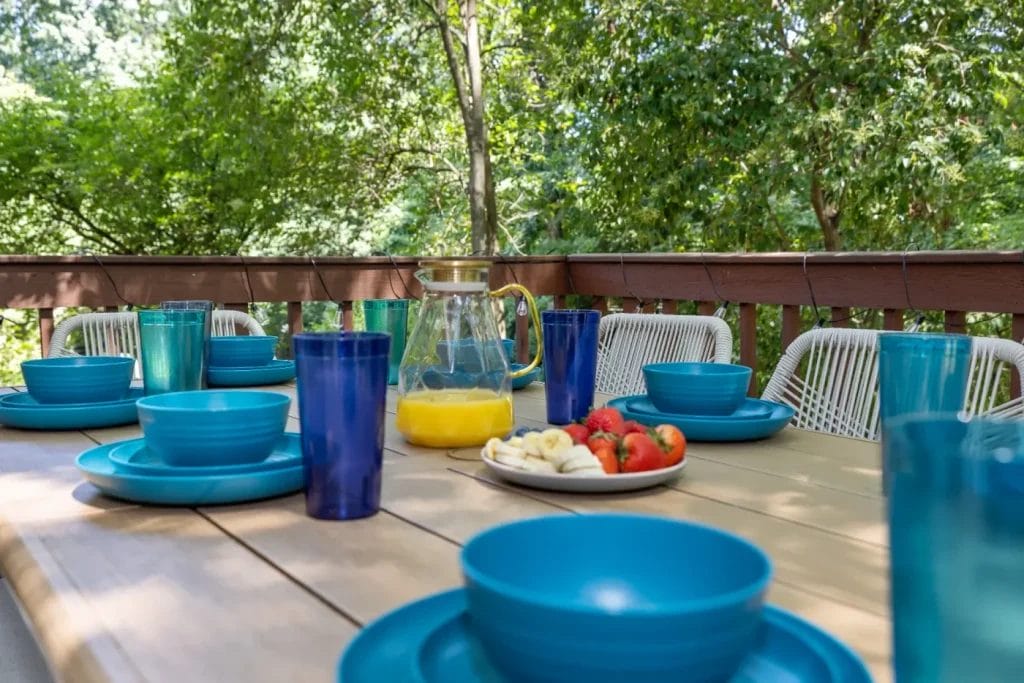 A wooden outdoor table set with blue dishes, blue cups, and a pitcher of orange juice. A plate of strawberries and sliced bananas is also on the table. Green trees and a wooden railing are in the background.