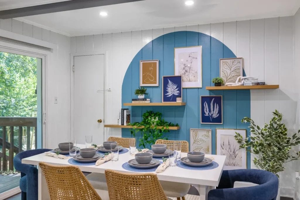 Modern dining room with a white table set for six, blue placemats, woven chairs, and blue curved accent wall featuring framed art, plants, and wooden shelves. Large window reveals greenery outside.