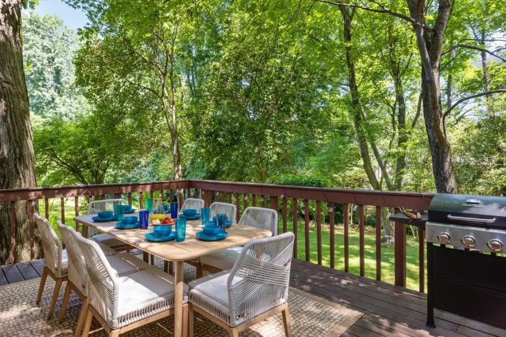 A wooden deck with a dining table set for six with blue dishes and glasses, white chairs, and a gas grill. The deck overlooks a lush, green, tree-filled backyard on a sunny day.